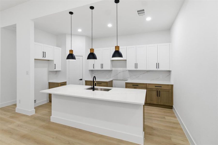 Kitchen with a kitchen island with sink, hanging light fixtures, white cabinetry, decorative backsplash, and light stone counters