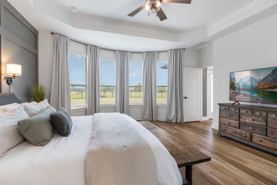 Spacious bedroom with large windows offering ample natural light, hardwood floors, and neutral tones. Features a ceiling fan, wall sconce, and a dresser with a mounted TV.