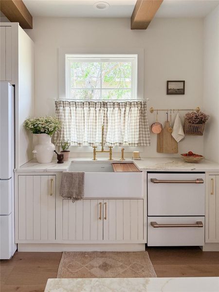 Kitchen featuring a white farmhouse sink with unlacquered brass faucet, white paneled cabinetry with brass hardware, double-drawer Forno dishwasher, and a window above the sink Kitchen featuring a white farmhouse sink with unlacquered brass faucet, white paneled cabinetry with brass hardware, double-drawer Forno dishwasher, and a window above the sink