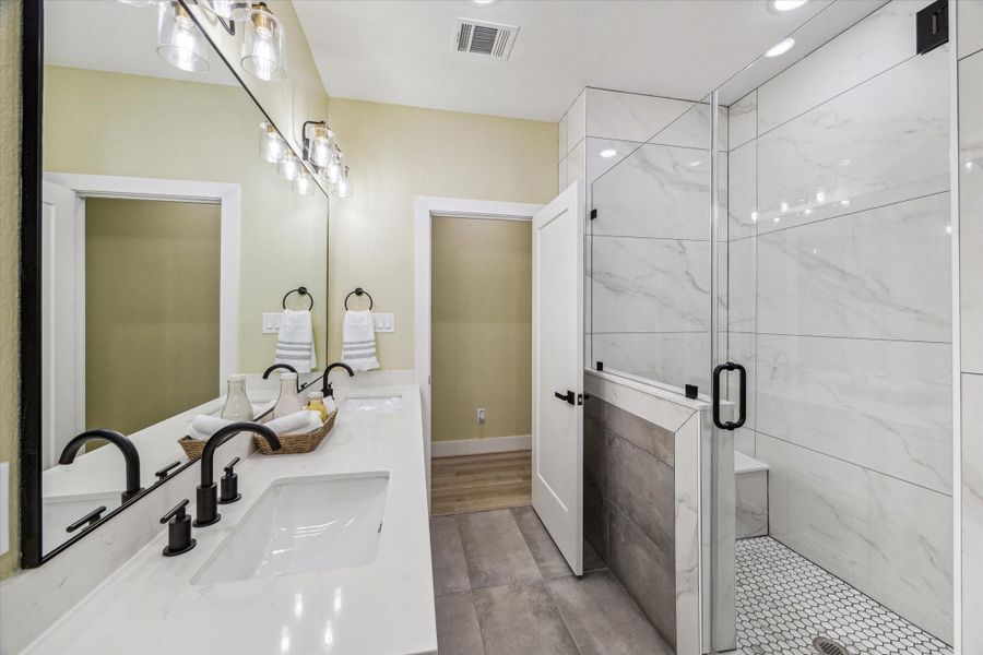 This modern bathroom features a double vanity with sleek black fixtures, a large mirror, and elegant lighting. The spacious walk-in shower is accented with marble-style tiles that continue uniquely on the wall and a glass door, adding a touch of luxury.