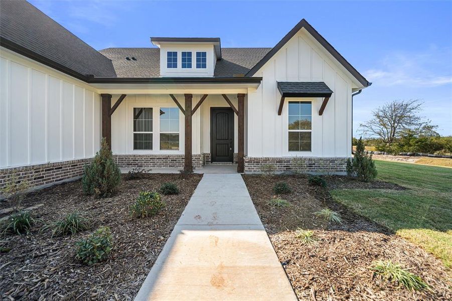 Entrance to property with board and batten siding, covered porch, a shingled roof, brick siding, and a yard