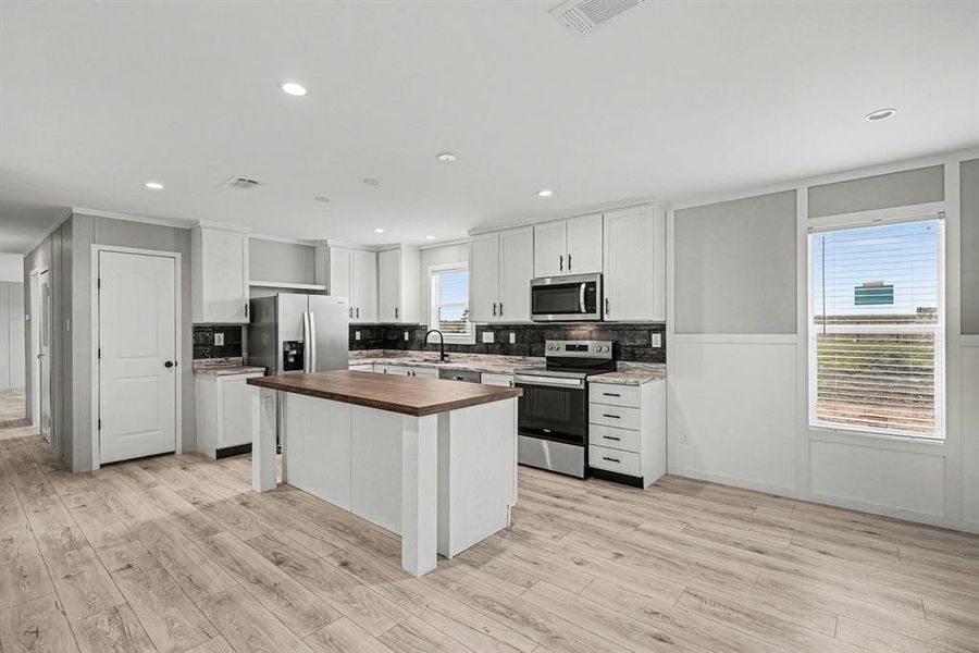 Well-appointed kitchen featuring a large central island, room for bar stools, focal-point decorative shelf above refrigerator, stainless steel appliances, white cabinetry, upgrade tile backsplash, and wood-look flooring. Door is to large walk-in pantry.