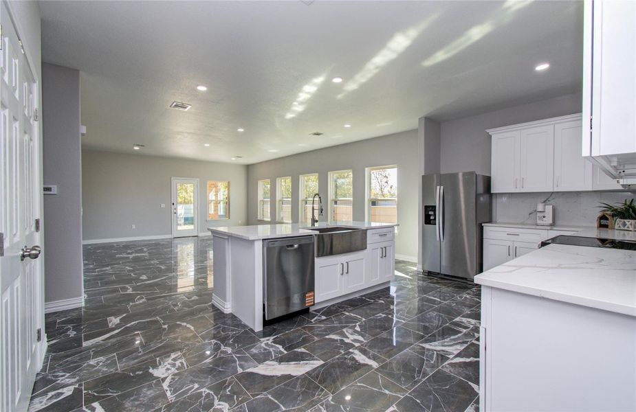 Spacious and modern open-concept kitchen with sleek marble floors, stainless steel appliances, and ample natural light from large windows. The central island offers additional workspace and storage, while the white cabinetry provides a clean, contemporary look.