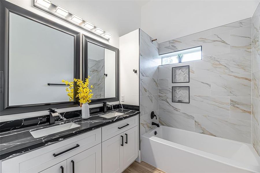 Modern secondary bathroom featuring a sleek double vanity with black marble countertop, large mirrors with bright overhead lighting, white cabinetry, and a full bathtub with elegant tile surround.