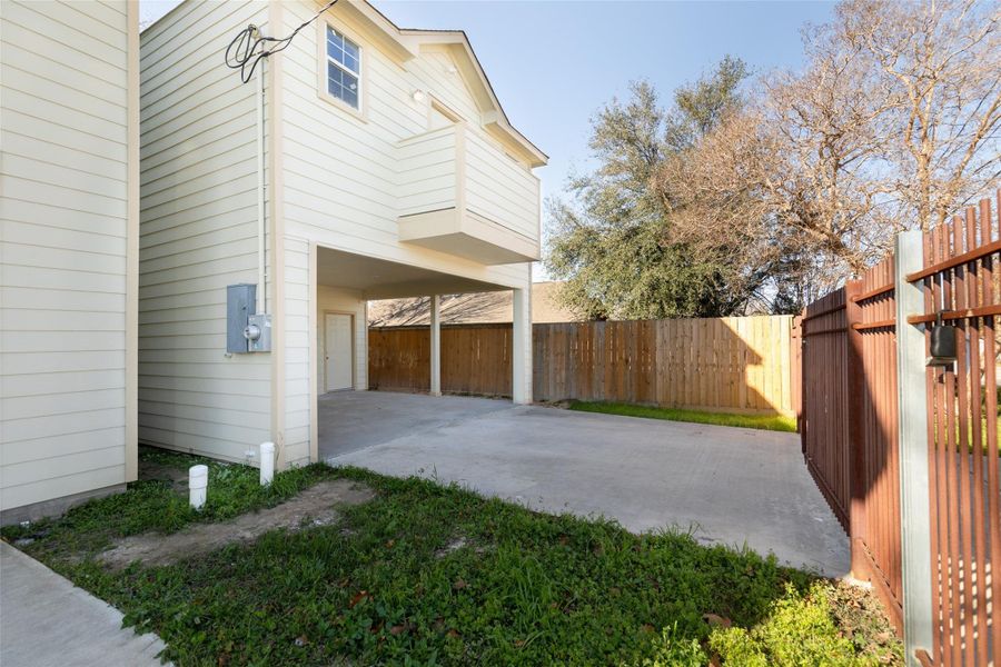 This photo shows a covered carport area attached to a house with beige siding. It opens onto a concrete driveway, enclosed by a wooden fence and a metal gate. There’s some grass and a few trees visible, offering a sense of privacy. This photo shows a covered carport area attached to a house with beige siding. It opens onto a concrete driveway, enclosed by a wooden fence and a metal gate. There’s some grass and a few trees visible, offering a sense of privacy.