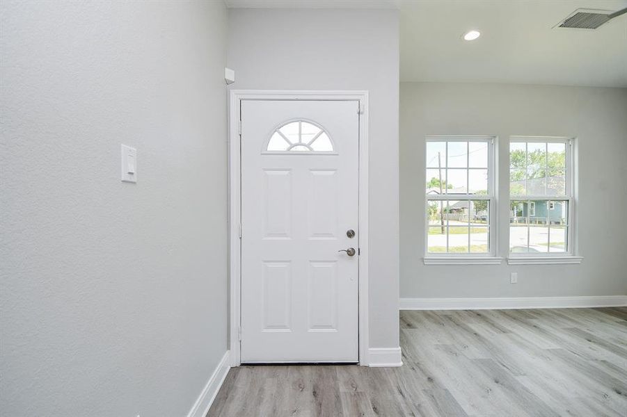 This photo shows a bright, modern entryway with a white front door featuring an arched window, complemented by a large double window that allows for ample natural light. The space has light grey walls and wood-look flooring, creating a clean and welcoming atmosphere.