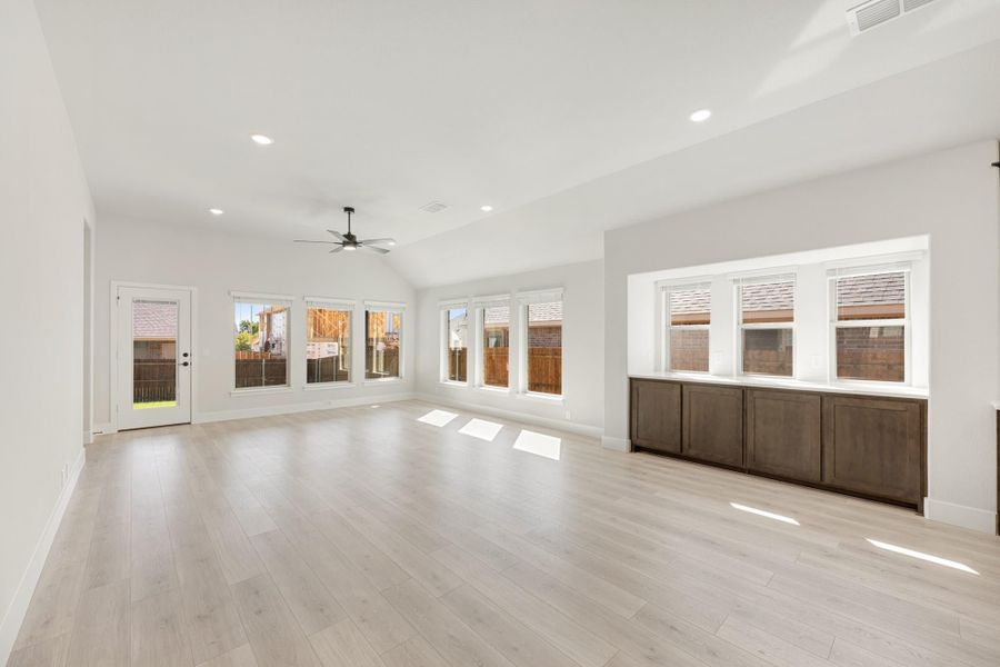 Open living room with vaulted ceiling, ceiling fan, light wood floors, and large windows with backyard views
