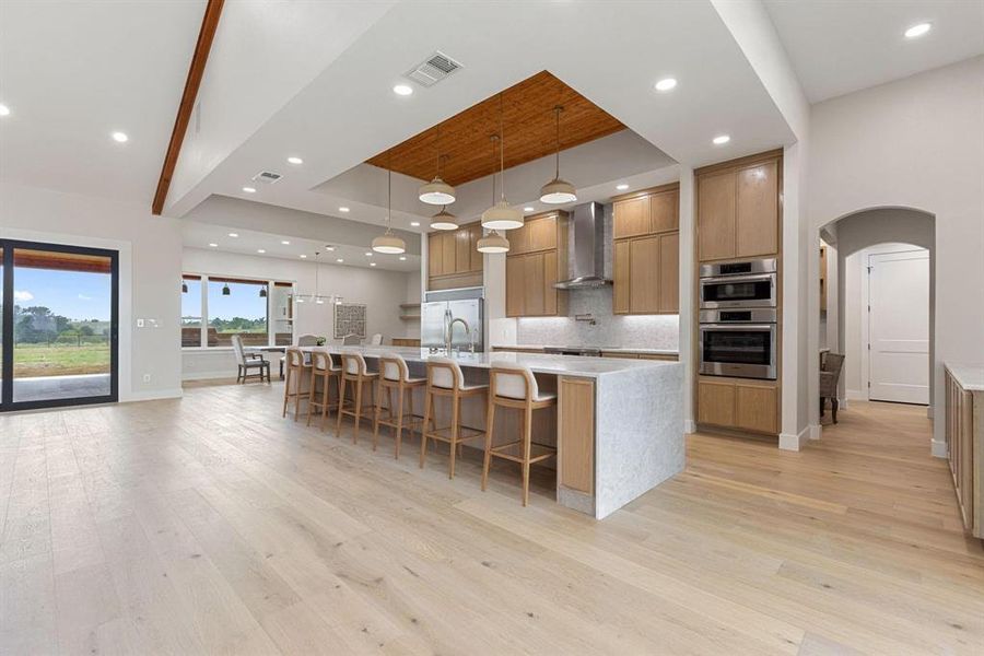 Kitchen featuring a tray ceiling, arched walkways,
light stone countertops, a large island with sink,
and hanging light fixtures Kitchen featuring a tray ceiling, arched walkways,
light stone countertops, a large island with sink,
and hanging light fixtures