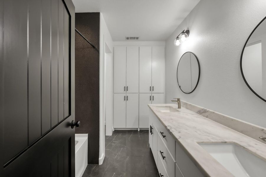 Bathroom #2 features a sleek design with a dual-sink marble countertop, modern black fixtures, circular mirrors, and ample storage with white cabinetry. The dark tile floor and textured wall add a touch of elegance.
