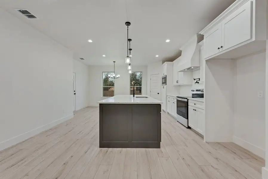 Kitchen featuring white cabinets, electric stove, custom exhaust hood, decorative light fixtures, and a center island with sink