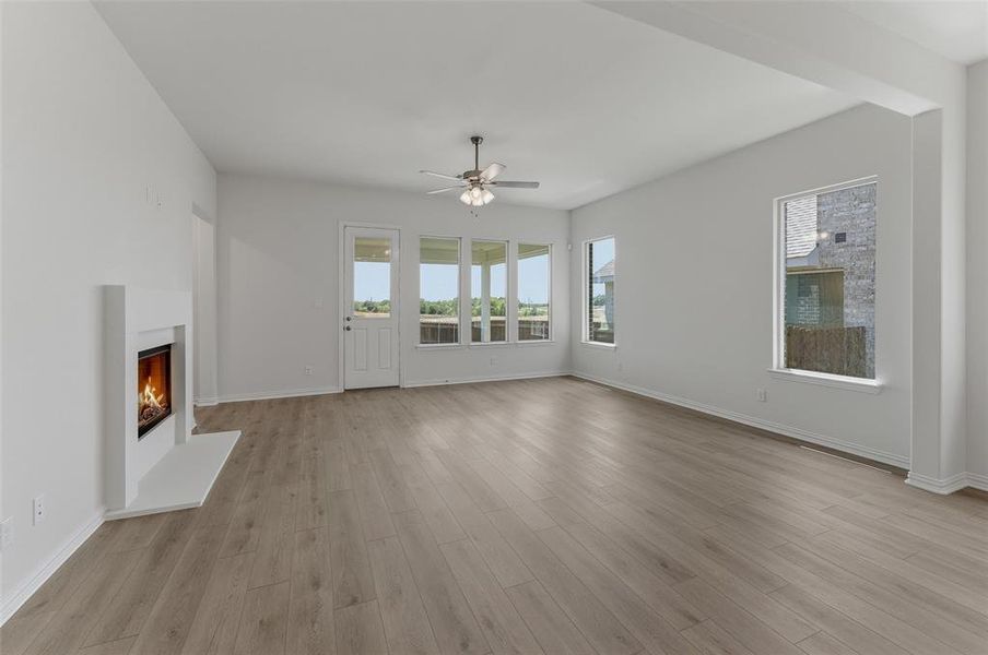 Spacious living area featuring light wood-finish flooring, a modern white fireplace, multiple windows with exterior views, and a ceiling fan Spacious living area featuring light wood-finish flooring, a modern white fireplace, multiple windows with exterior views, and a ceiling fan