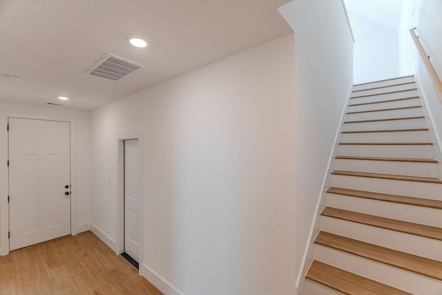 This photo shows a modern, bright entryway featuring wood flooring, a simple white door, and a staircase with wood-accented steps leading upwards. The space is well-lit with recessed lighting, creating a welcoming atmosphere.Photo Disclaimer: Photos are representative of the community and may depict a similar floor plan, model home, or previously completed residence. This photo shows a modern, bright entryway featuring wood flooring, a simple white door, and a staircase with wood-accented steps leading upwards. The space is well-lit with recessed lighting, creating a welcoming atmosphere.Photo Disclaimer: Photos are representative of the community and may depict a similar floor plan, model home, or previously completed residence.