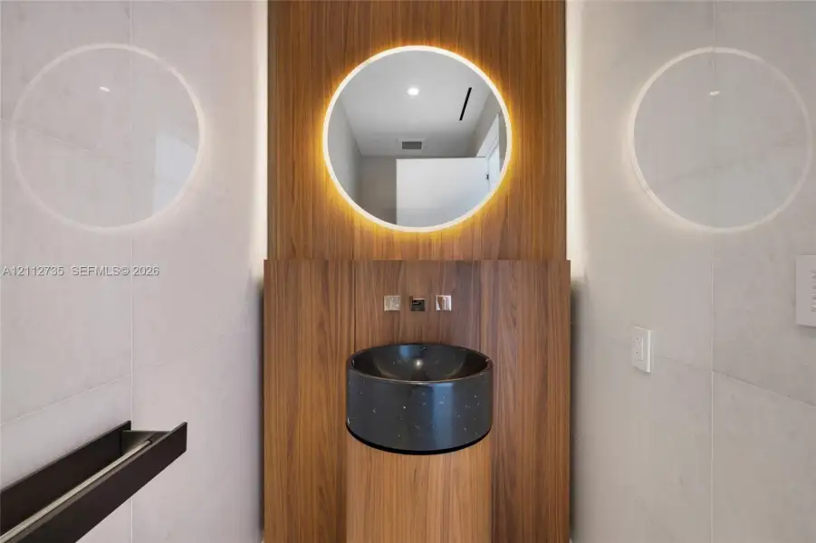 Wall-mounted wood-finish paneling, circular backlit mirror, cylindrical wood-finish pedestal vanity, black stone vessel sink, and wall-mounted metallic fixtures