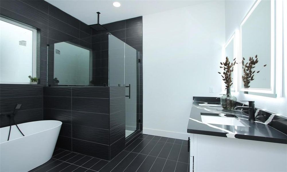 This primary bathroom features oversized black tiles, a spacious glass enclosed custom shower, a freestanding white tub, and a stylish dual-sink vanity with illuminated mirrors.