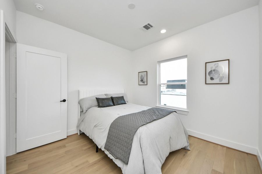 This is a bright, modern bedroom featuring a comfortable bed with neutral bedding. The room has light wood flooring, white walls, a window providing natural light, and simple wall art, creating a clean and inviting atmosphere. This is a bright, modern bedroom featuring a comfortable bed with neutral bedding. The room has light wood flooring, white walls, a window providing natural light, and simple wall art, creating a clean and inviting atmosphere.