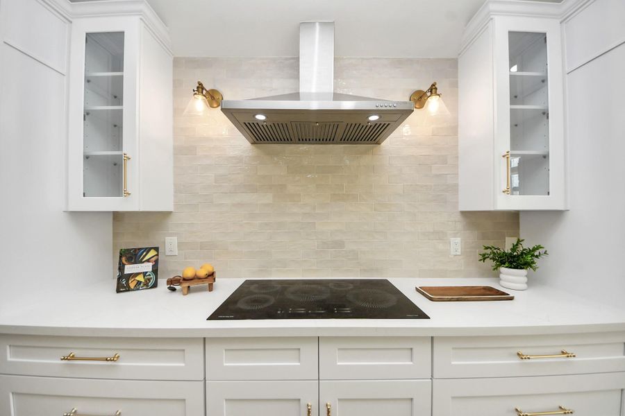 A beautifully curated kitchen where modern white cabinetry with glass accents meets warm brass hardware and soft, ambient lighting. The sleek induction cooktop and stainless steel range hood offer a streamlined look, while a beige subway tile backsplash brings just the right touch of warmth and texture. A beautifully curated kitchen where modern white cabinetry with glass accents meets warm brass hardware and soft, ambient lighting. The sleek induction cooktop and stainless steel range hood offer a streamlined look, while a beige subway tile backsplash brings just the right touch of warmth and texture.