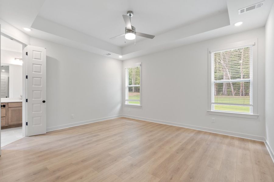 Tray ceiling with recessed lighting, light wood-finish flooring, two windows with blinds, modern ceiling fan with light, and white paneled door with matte black hardware Tray ceiling with recessed lighting, light wood-finish flooring, two windows with blinds, modern ceiling fan with light, and white paneled door with matte black hardware