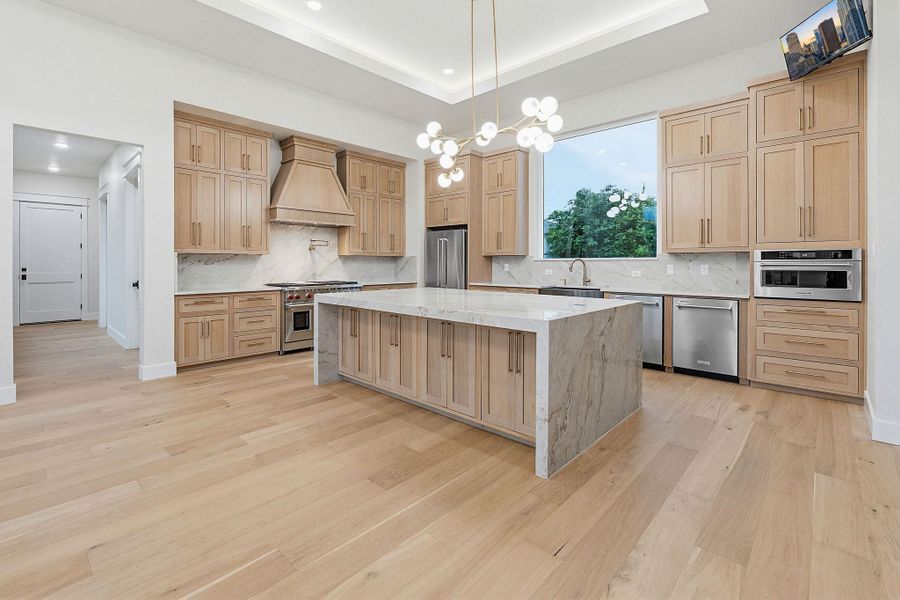 Gourmet kitchen featuring a waterfall island, light wood-finish cabinetry, stainless steel appliances, and a large picture window