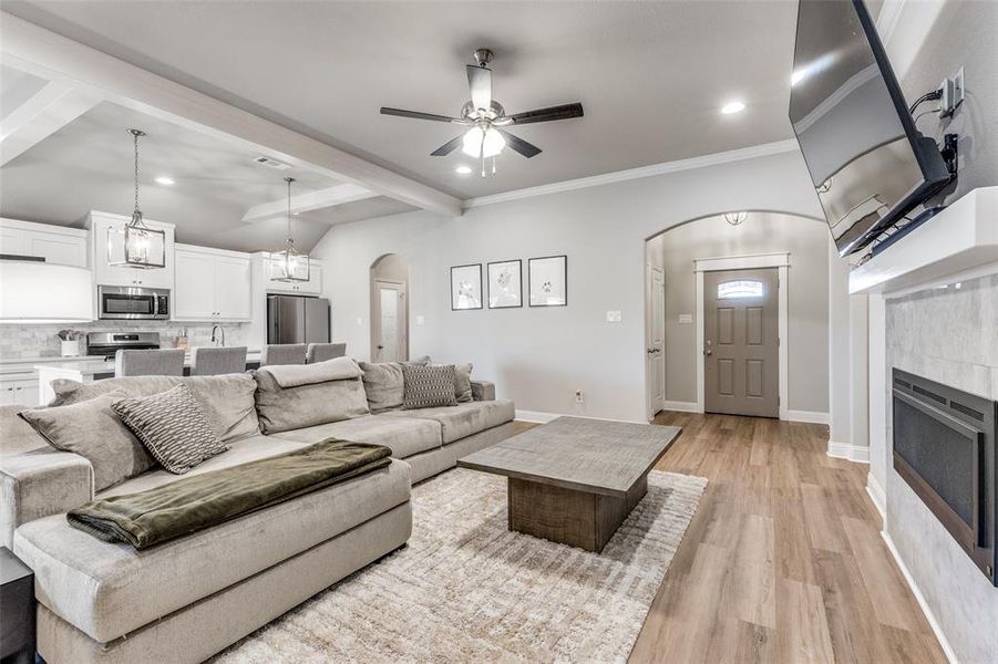 Living area with arched walkways, beam ceiling, a tile fireplace, light wood-style flooring, and ceiling fan Living area with arched walkways, beam ceiling, a tile fireplace, light wood-style flooring, and ceiling fan