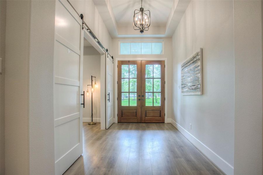 This welcoming entryway features double wooden doors with glass panels, high ceilings, a modern chandelier, and light wood flooring. A barn-style sliding door and contemporary artwork add style and functionality. Natural light enhances the bright, airy ambiance.