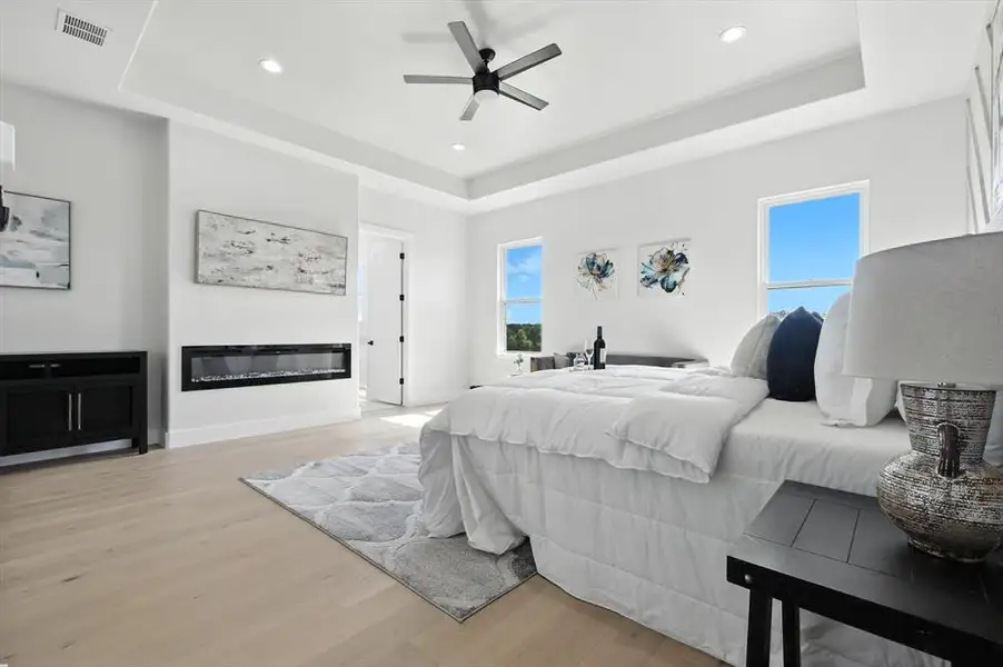 Bedroom with a tray ceiling, light wood finished floors, a glass covered fireplace, recessed lighting, and a ceiling fan Bedroom with a tray ceiling, light wood finished floors, a glass covered fireplace, recessed lighting, and a ceiling fan