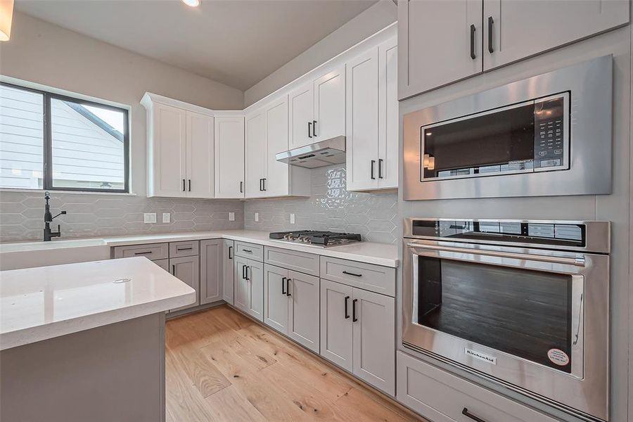 Discover a modern kitchen with sleek white cabinetry, stainless steel appliances, and a chic hexagonal tile backsplash. Enjoy abundant natural light from a large window and a spacious layout featuring a built-in oven and microwave.