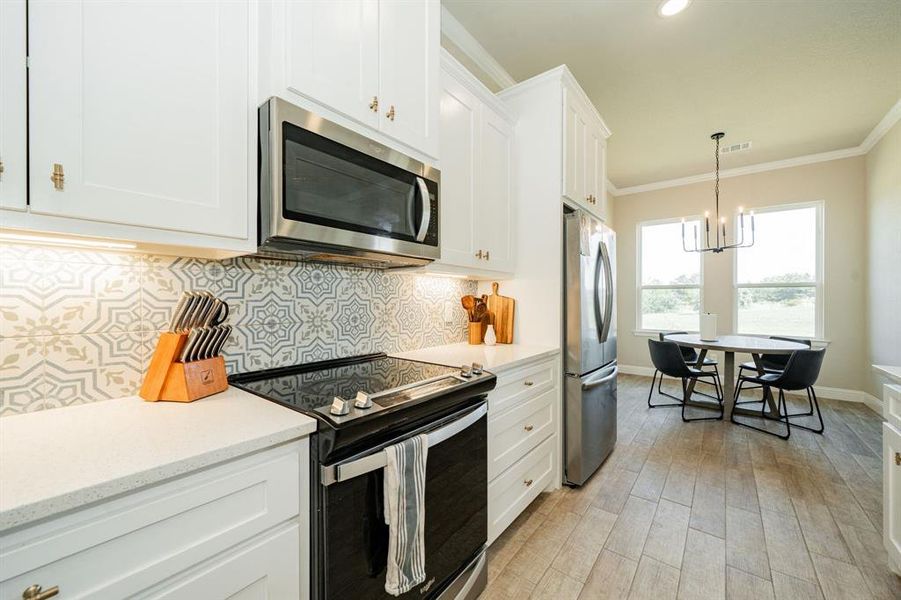 Kitchen featuring stainless steel appliances, backsplash, ornamental molding, white cabinetry, and decorative light fixtures Kitchen featuring stainless steel appliances, backsplash, ornamental molding, white cabinetry, and decorative light fixtures