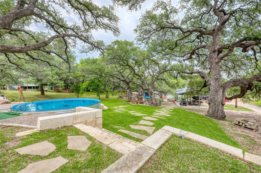 Expansive grassy yard featuring mature trees, a stock tank pool, stone retaining wall, and flagstone pathway