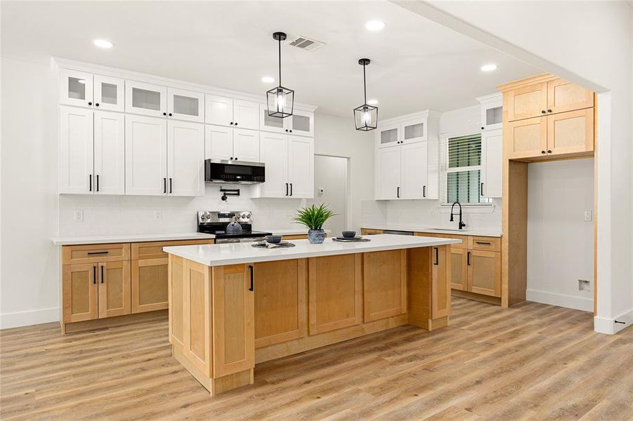 This modern kitchen features a spacious island with light wood cabinetry, contrasting with white upper cabinets. It includes sleek black fixtures, pendant lighting, and a bright, open layout with a stylish wood floor. Ideal for cooking and entertaining.