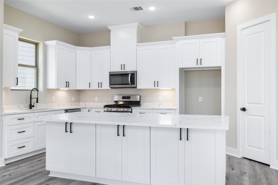 The kitchen, complete with modern finishes, showcases sleek white cabinetry, an expansive quartz countertop island, stainless steel appliances, and elegant lighting — a welcoming space designed for gathering, cooking, and making memories.