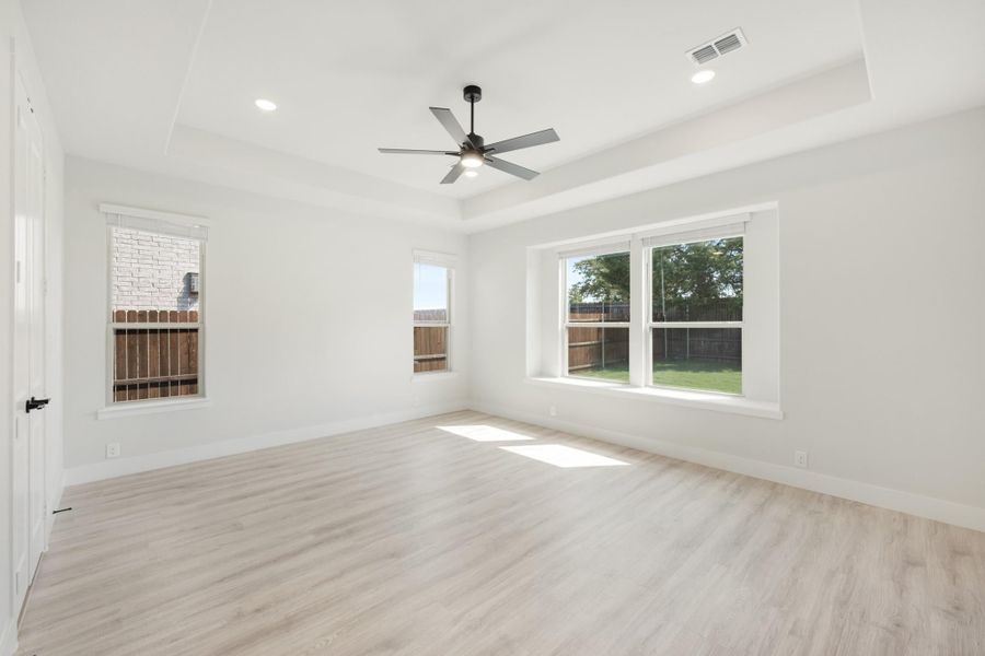 Empty room with tray ceiling, ceiling fan, light wood floors, and large windows overlooking backyard