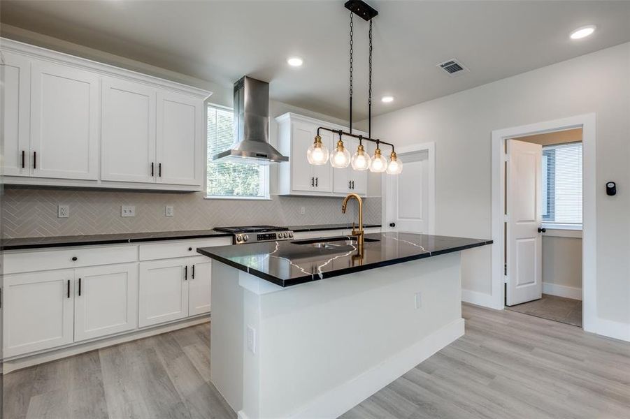 Kitchen with an island with sink, wall chimney range hood, decorative backsplash, hanging light fixtures, and white cabinets