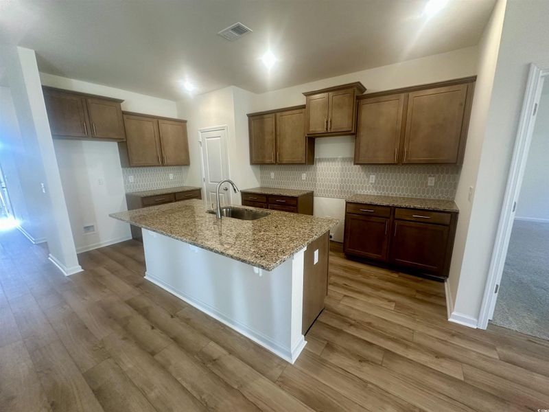 Kitchen featuring granite counters, brown cabinetry, an island with sink, beautiful backsplash, and a breakfast bar area