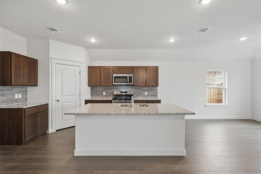 Kitchen with light stone countertops, dark wood-style floors, stainless steel appliances, a center island with sink, and backsplash Kitchen with light stone countertops, dark wood-style floors, stainless steel appliances, a center island with sink, and backsplash