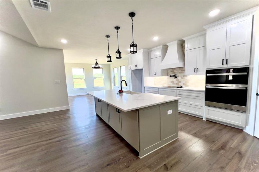 Kitchen featuring backsplash, custom exhaust hood, a center island with sink, white cabinets, and dark floors