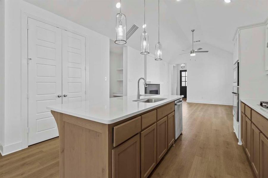 Kitchen featuring decorative light fixtures, a center island with sink, lofted ceiling, light wood-type flooring, and brown cabinetry Kitchen featuring decorative light fixtures, a center island with sink, lofted ceiling, light wood-type flooring, and brown cabinetry