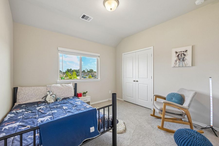 Charming bedroom with soft neutral walls, plush carpeting, and a cozy bed under a bright window offering natural light. A modern rocking chair complements the space, providing a relaxing retreat.