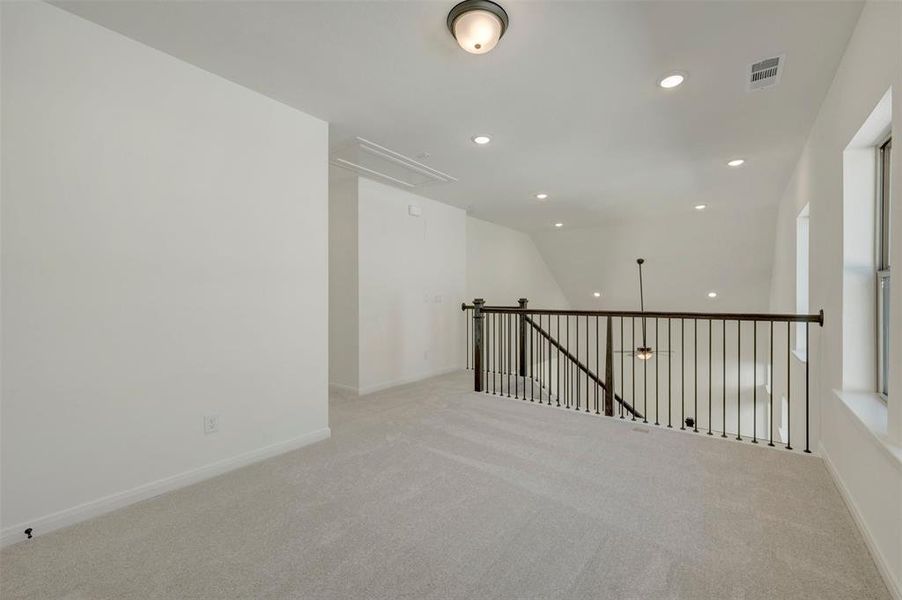 Spacious loft area featuring light gray carpeting, white walls, multiple recessed lights, a ceiling-mounted light fixture, and an iron baluster railing overlooking the floor below Spacious loft area featuring light gray carpeting, white walls, multiple recessed lights, a ceiling-mounted light fixture, and an iron baluster railing overlooking the floor below