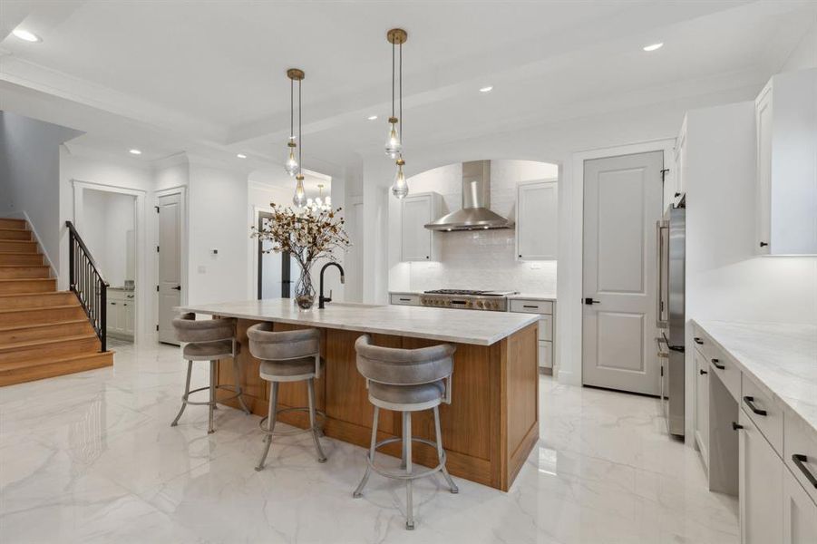 Kitchen featuring tasteful backsplash, decorative light fixtures, a center island with sink, wall chimney range hood, and white cabinetry