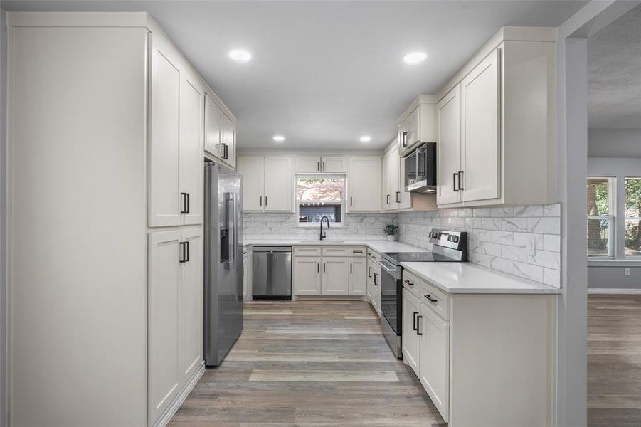 Galley kitchen featuring light shaker cabinetry, light countertops, subway tile backsplash, stainless steel appliances, and wood-finish flooring Galley kitchen featuring light shaker cabinetry, light countertops, subway tile backsplash, stainless steel appliances, and wood-finish flooring