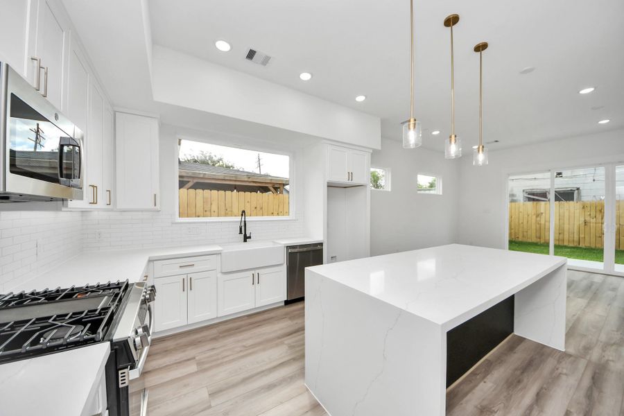 This modern kitchen features sleek white cabinetry, a large island with elegant pendant lighting, and stainless steel appliances. Ample natural light flows through large windows and glass doors, enhancing the open and airy feel.