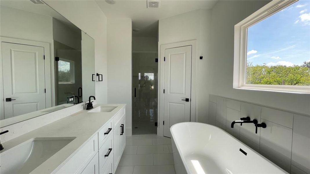 Private ensuite featuring a double vanity with ample storage, sleek black fixtures, a large mirror, and a freestanding tub near a window with a pleasant view.