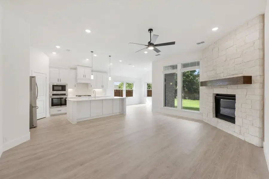 Open-concept living area with stone fireplace, light wood floors, and white kitchen with island and stainless appliances