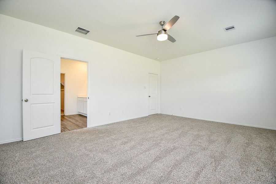 This spacious bedroom features neutral carpeting, white walls, and a ceiling fan. It offers ample natural light and includes a walk-in closet and an en-suite bathroom with wood flooring.