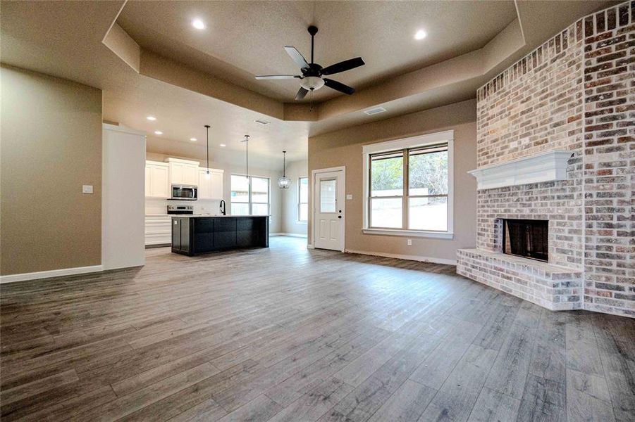 Unfurnished living room with ceiling fan, wood-type flooring, a tray ceiling, and a brick fireplace