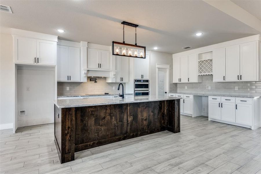 Kitchen featuring a kitchen island with sink, decorative light fixtures, white cabinetry, stainless steel double oven, and light stone countertops