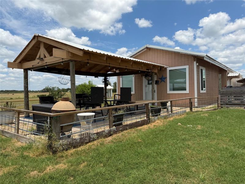 An inviting covered patio adjoins a charming home, overlooking open fields under a bright sky.