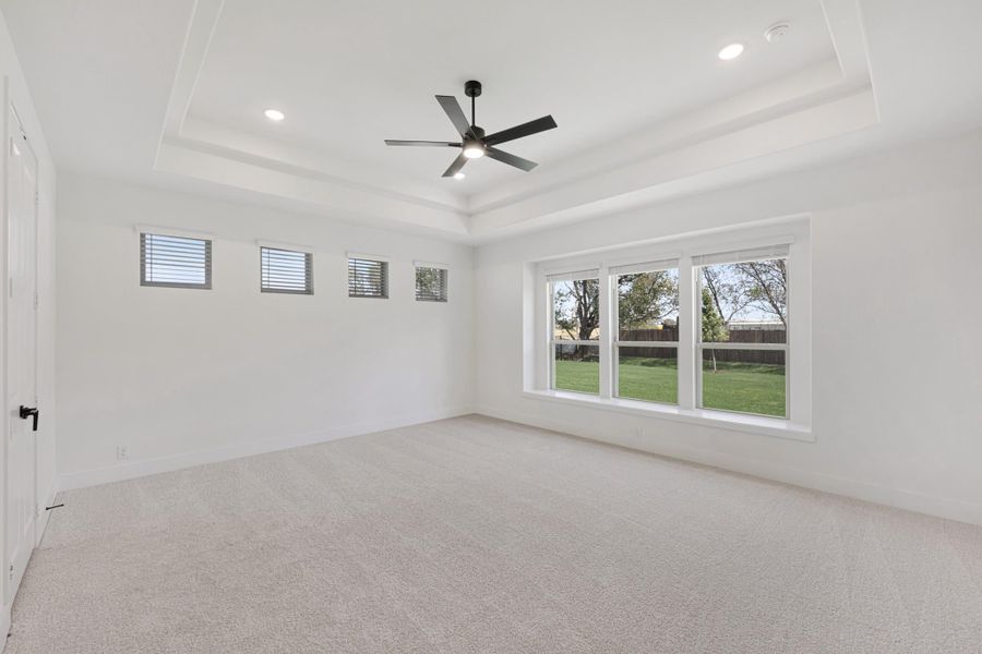 Empty bedroom with tray ceiling, black ceiling fan, carpet flooring, and large window overlooking backyard