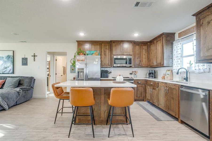 Kitchen with a center island, brown wood cabinetry, backsplash, open floor plan, and appliances with stainless steel finishes Kitchen with a center island, brown wood cabinetry, backsplash, open floor plan, and appliances with stainless steel finishes