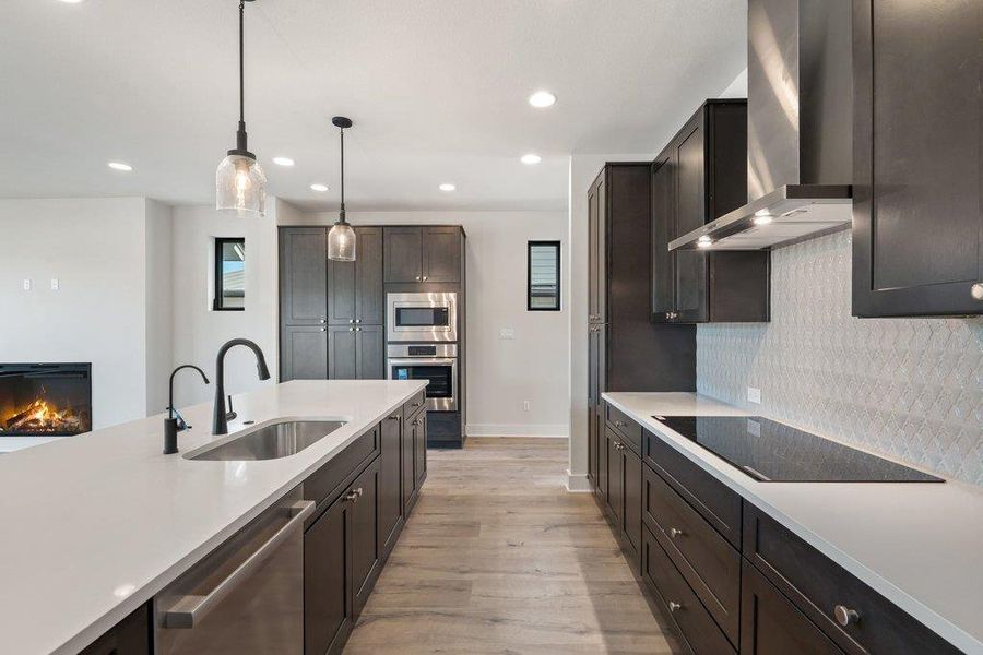 Kitchen featuring wall chimney range hood, appliances with stainless steel finishes, decorative light fixtures, light wood finished floors, and a lit fireplace